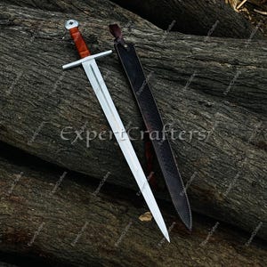 May include: A medieval-style sword with a silver blade and a brown handle, resting on a wooden surface. The sword is accompanied by a black leather sheath. The image has the text "Expert Crafters".