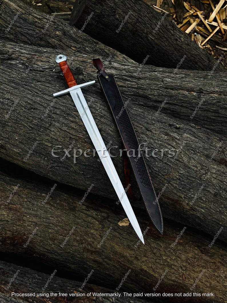May include: A medieval-style sword with a silver blade and a brown handle, resting on a wooden surface. The sword is accompanied by a black leather sheath. The image has the text "Expert Crafters".