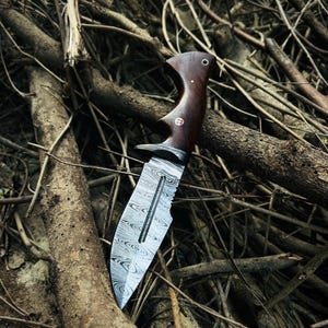 May include: A Damascus steel knife with a dark wood handle. The blade has a unique pattern and a serrated edge. The handle features a curved design with a small, decorative emblem. The knife is set against a backdrop of twigs and branches.