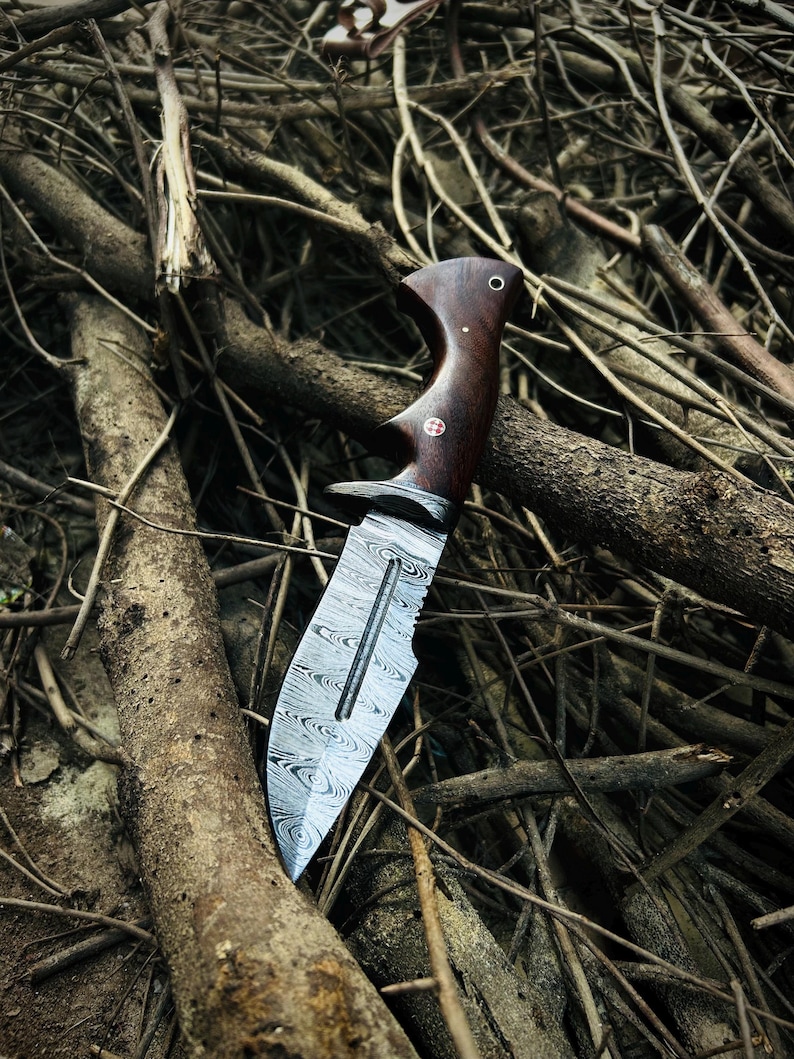 May include: A Damascus steel knife with a dark wood handle. The blade has a unique pattern and a serrated edge. The handle features a curved design with a small, decorative emblem. The knife is set against a backdrop of twigs and branches.