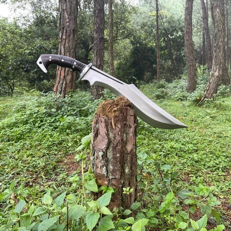 May include: A large, silver-colored knife with a curved blade and a dark wooden handle. The knife is resting on a tree stump in a forest setting, surrounded by green foliage and trees. The blade has a sharp edge and a decorative design.