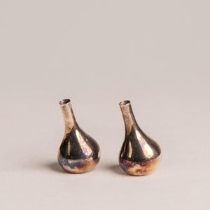 Può includere: Due piccoli vasi a bocciolo color argento con corpo a forma di pera e collo sottile. I vasi hanno una lucentezza metallica con sfumature di blu e oro. Lo sfondo è grigio chiaro.