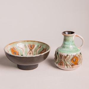 May include: A ceramic bowl and pitcher set. The bowl is dark brown with a light green and orange patterned interior. The pitcher is light green with a brown handle and base, featuring an orange and brown design.