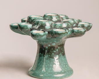 Könnte beinhalten: Ein teal-farbener Kerzenhalter aus Keramik mit mehreren kleinen, becherförmigen Haltern. Der Kerzenhalter hat ein baumartiges Design mit breitem Sockel und einer strukturierten, gesprenkelten Oberfläche. Die teal-Farbe geht in ein dunkleres Grün über.