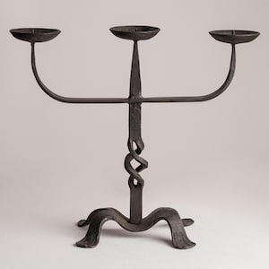Könnte beinhalten: Ein schmiedeeiserner Kerzenständer mit drei Kerzenhaltern. Der dunkle Metall-Kerzenständer hat eine gedrehte Mittelstütze und gebogene Arme. Der Sockel hat ein dekoratives, geschwungenes Design. Die Kerzenhalter sind kreisförmig und flach.