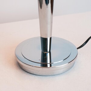 May include: A polished chrome lamp base featuring a circular, flat foundation and a tapered, vertical cylindrical stem. A thin black electrical cord extends from the side of the base, which has a smooth, reflective metallic surface throughout.