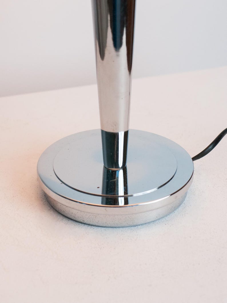 May include: A polished chrome lamp base featuring a circular, flat foundation and a tapered, vertical cylindrical stem. A thin black electrical cord extends from the side of the base, which has a smooth, reflective metallic surface throughout.