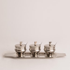 Peut inclure: Un ensemble de trois pots à condiments en acier inoxydable avec couvercles et petites cuillères, présentés sur un plateau rectangulaire assorti. Les pots ont un design classique et vintage, idéal pour servir du sucre, du sel ou des épices.