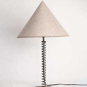 May include: A table lamp featuring a conical beige fabric lampshade and a base constructed from a dark metal coil spring attached to a circular metal plate. A black electrical cord extends from the base of the lamp to provide power.