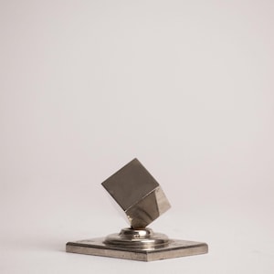 Op de afbeelding: Een zilverkleurig metalen sculptuur bestaande uit een kubus die op één hoek balanceert bovenop een platte, vierkante voet. Het object heeft een glad, reflecterend oppervlak en een metalen afwerking, waarbij de kubus schuin omhoog staat.