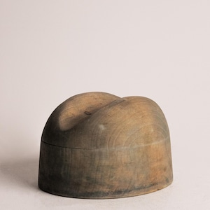 May include: A vintage wooden hat block with a rounded, domed shape. The hat block is made of aged wood with a natural finish, showing variations in color from light brown to darker, weathered tones. The top has a slight indentation.