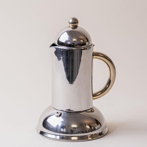 May include: A shiny, stainless steel stovetop espresso maker with a gold-colored handle. The coffee pot has a rounded base and a domed lid with a small, round knob. The polished metal reflects the surrounding environment.