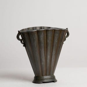 Può includere: Un vaso in metallo grigio scuro con un corpo scanalato a forma di ventaglio e un bordo largo e smerlato. Il recipiente presenta due piccole maniglie decorative a forma di voluta sui lati opposti e poggia su una base circolare leggermente rialzata.
