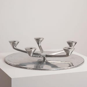 May include: A silver-toned metal candelabra featuring a flat, circular base and four upward-curving arms, each ending in a conical candle holder. The object has a polished, reflective surface and a low-profile, symmetrical design.