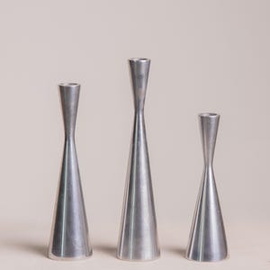 May include: Three silver candle holders of different heights. Each has a conical base leading to a narrow, cylindrical top. The candle holders are arranged in a row against a neutral background.
