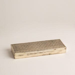 Op de afbeelding: Een rechthoekige doos met een metallic afwerking en een verweerde uitstraling. Het deksel heeft een herhalend geometrisch patroon van kleine, donkere, in elkaar grijpende lijnen op een lichtere achtergrond, terwijl de zijkanten een versleten zilveren textuur tonen.
