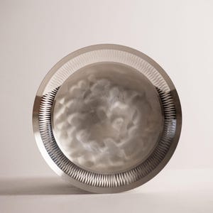 Könnte beinhalten: Ein kreisförmiger silberfarbener Metallteller mit einer glatten, reflektierenden Mitte und einem subtilen, wolkenartigen Texturmuster. Der äußere Rand ist durch eine breite, polierte Kante definiert, die mit einem feinen, gezackten Muster verziert ist.