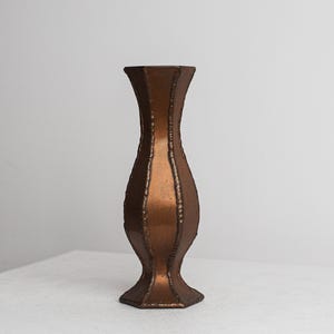 Puede incluir: Un jarrón alto de color bronce con un diseño texturizado único. El jarrón tiene una parte superior acampanada y una base ancha, con una serie de crestas verticales a lo largo del cuerpo. El acabado metálico le da un aspecto vintage.