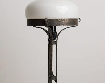 Peut inclure: Une lampe de table dotée d'une base en métal martelé sombre et texturé avec une plaque inférieure hexagonale et des supports incurvés. La lampe est surmontée d'un abat-jour en dôme de verre blanc opaque et lisse, avec un cordon électrique noir.
