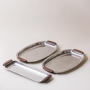 Pode incluir: Três travessas de servir em aço inoxidável com pegas de madeira. As travessas são ovais e retangulares, com um acabamento em metal escovado. As pegas de madeira são de cor castanho escuro. As travessas estão dispostas numa superfície branca.