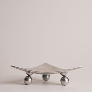 Puede incluir: Un plato decorativo moderno de color plateado, de forma cuadrada y bordes curvos. El plato está sostenido por tres pies esféricos, creando un diseño minimalista y elegante. El metal tiene un acabado cepillado.