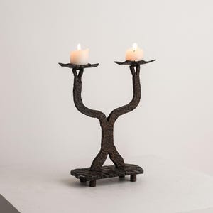 May include: A dark brown wrought iron candle holder with two lit candles. The candle holder has a textured, organic design with two arms holding the candles. The base is rectangular with four small feet. The candles are white.