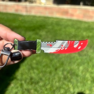 Puede incluir: Un llavero con forma de cuchillo con una hoja plateada, mango negro y detalles rojos. El llavero tiene un cierre plateado y un borde verde. La hoja tiene un diseño de salpicaduras de sangre.