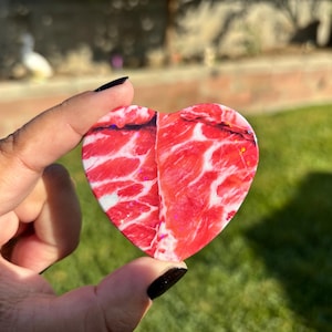 Puede incluir: Un objeto en forma de corazón con un diseño veteado rojo y blanco, que recuerda a la carne cruda. El objeto se sostiene contra un fondo verde borroso. La mano que lo sostiene tiene esmalte de uñas negro. El diseño tiene vetas rojas y blancas.