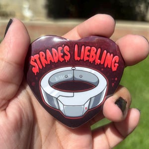 Puede incluir: Botón en forma de corazón con el texto "STRADE'S LIEBLING" en letras rojas estilizadas. El botón presenta un diseño blanco y gris de un collar con la palabra "BUDDY" y un símbolo de corazón. El fondo es rojo oscuro.