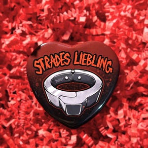 May include: A heart-shaped pin featuring a dark red and black gradient background with the text STRADES LIEBLING in orange letters. The center displays a graphic illustration of a silver metallic collar with the word BUDDY and a heart symbol.