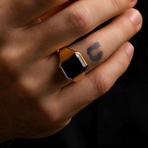Puede incluir: Un anillo dorado con una piedra negra rectangular. El anillo está en un dedo. El anillo tiene un acabado pulido. El fondo es oscuro.