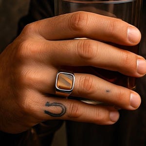 May include: A silver ring with a square, tiger's eye stone. The ring is on a hand holding a glass of amber liquid. The background is a dark brown.