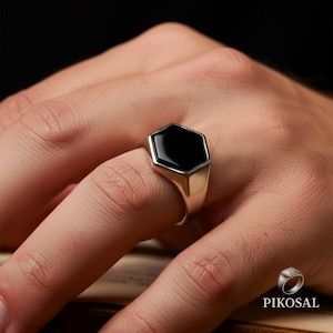 Puede incluir: Un anillo de plata con una piedra de ónix negra hexagonal. El anillo tiene un acabado pulido y se muestra en un dedo. El anillo es un estilo de sello clásico.