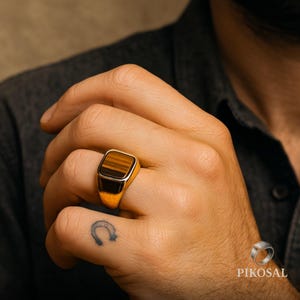 May include: A gold signet ring featuring a rectangular tiger's eye stone. The ring has a polished gold band and a dark brown stone with brown and black stripes. The ring is on a person's finger.