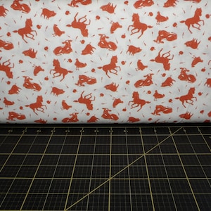 May include: White fabric with a repeating pattern of red horses and other animals. The fabric is folded in half and lying on a black cutting mat.