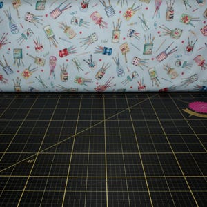 Peut inclure: Gros plan sur un tissu bleu avec un motif répété de pinceaux dans des pots. Le tissu est plié sur un tapis de coupe noir avec des lignes de quadrillage blanches.