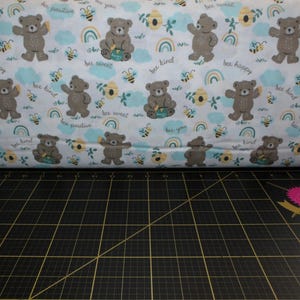 May include: A roll of fabric with a repeating pattern of brown teddy bears, bees, rainbows, and floral elements on a white background. The fabric includes the phrases "bee happy", "bee kind", and "bee positive".
