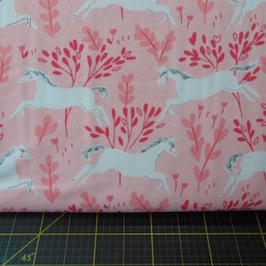 May include: Pink fabric with a white unicorn pattern. The unicorns are running through a field of pink flowers and leaves.