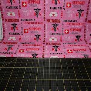 May include: Pink fabric with a repeating pattern of the words "Nurses", "RN", "Caring", "Emergency", "Encourage", "Service", "Support", "Care", "Save a Life", and "Listen". The pattern also includes a red cross and a caduceus symbol.