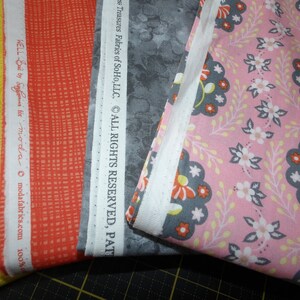 Puede incluir: Tres muestras de tela, una amarilla con un patrón de espiral, una naranja con un patrón de cuadrícula y una gris con un patrón floral blanco. El texto "Treasures Fabrics of SoHo, LLC. ALL RIGHTS RESERVED, PAT" está impreso en la tela gris. El texto "WELL Baby Sfor moda modafabrics.com 100%" está impreso en la tela naranja. El texto "Just Color by" está impreso en la tela amarilla.