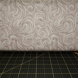 May include: A close-up of a beige fabric with a swirling pattern. The fabric is folded over a black cutting mat with a grid pattern.