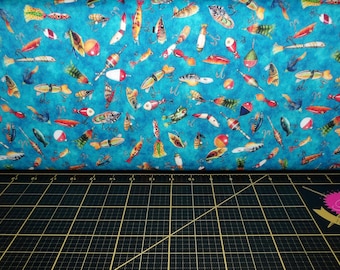 May include: A blue fabric with a repeating pattern of fishing lures and bobbers. The fabric is on a black cutting mat.