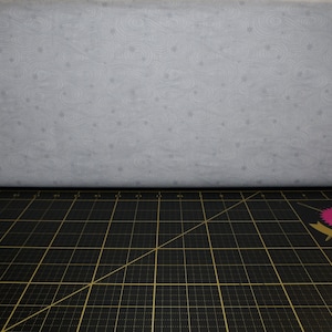 Puede incluir: Primer plano de una tela blanca con un sutil patrón de estrellas y remolinos. La tela está doblada sobre una estera de corte negra con un patrón de cuadrícula. Una pegatina rosa y dorada con el texto "Tula Handmade" es visible en la esquina inferior derecha.