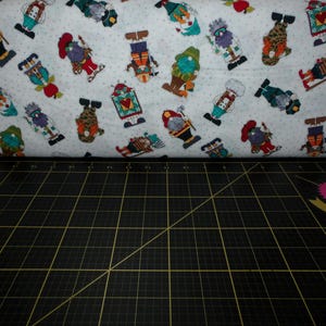 May include: White fabric with a repeating pattern of colorful gnomes. The gnomes are wearing different hats and holding various items. The fabric is folded in half and lying on a black cutting mat.