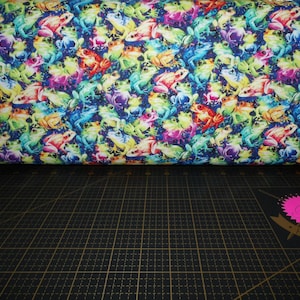 Puede incluir: Tela con un patrón repetido de ranas coloridas en tonos rojos, naranjas, amarillos, verdes, azules y morados. Las ranas están dispersas sobre un fondo azul oscuro. La tela está enrollada, con una superficie de cuadrícula negra en primer plano.