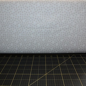 May include: A close-up of a white fabric with a subtle gray polka dot pattern. The fabric is lying on a black cutting mat.