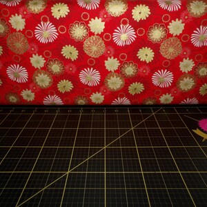 May include: Red fabric with a white and gold floral pattern. The fabric is folded over a black cutting mat with white grid lines.