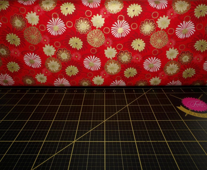 May include: Red fabric with a white and gold floral pattern. The fabric is folded over a black cutting mat with white grid lines.