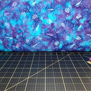 May include: A close-up of a blue and purple batik fabric with a white floral pattern. The fabric is lying on a black cutting mat with a ruler and a pink and gold label that says "Tula Pink" on it.