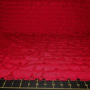 Könnte beinhalten: Nahaufnahme eines leuchtend roten Stoffes mit einem strukturierten, geschichteten Design. Der Stoff scheint gerafft oder gerüscht zu sein und erzeugt horizontale Texturbänder. Die Farbe ist ein tiefes, sattes Rot, und der Stoff hat einen leichten Glanz.